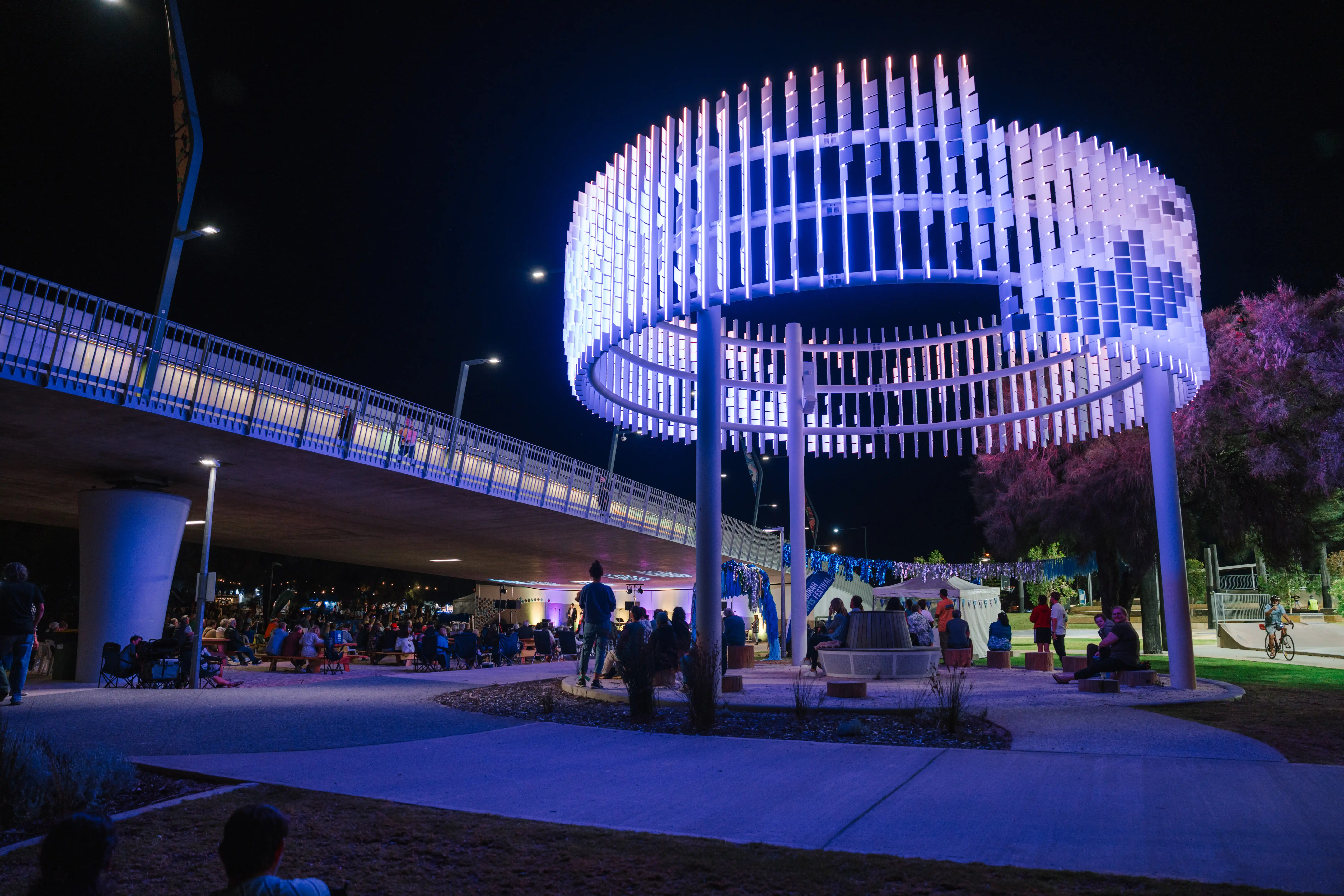 A circular light sculpture glowing blue at night with people gathered beneath it near a bridge.
